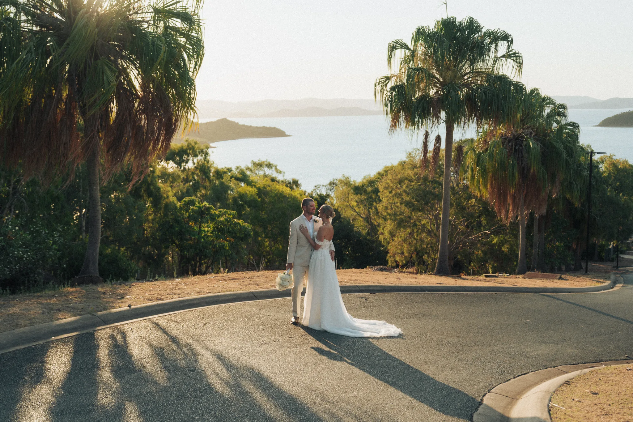 The bride walks a palm-lined path with long shadows across the ground